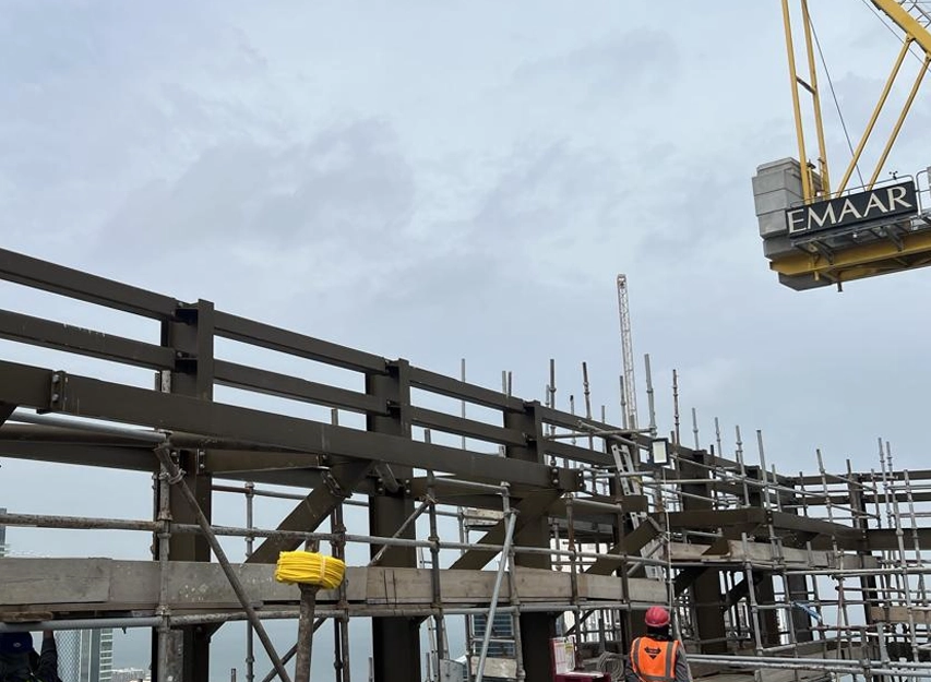 Steel frame and scaffolding on a high-rise rooftop with a worker in a hard hat and a tower crane above
