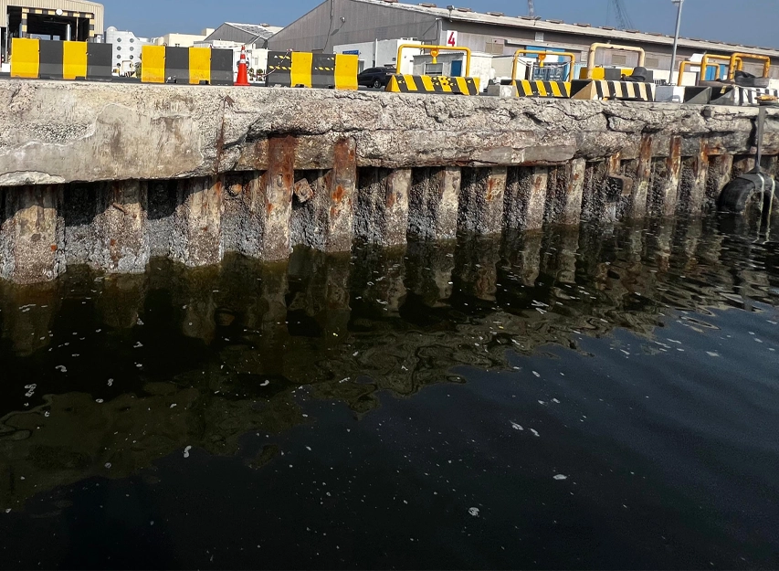 Corroded concrete quay wall with rust-stained, spalled piles above the waterline